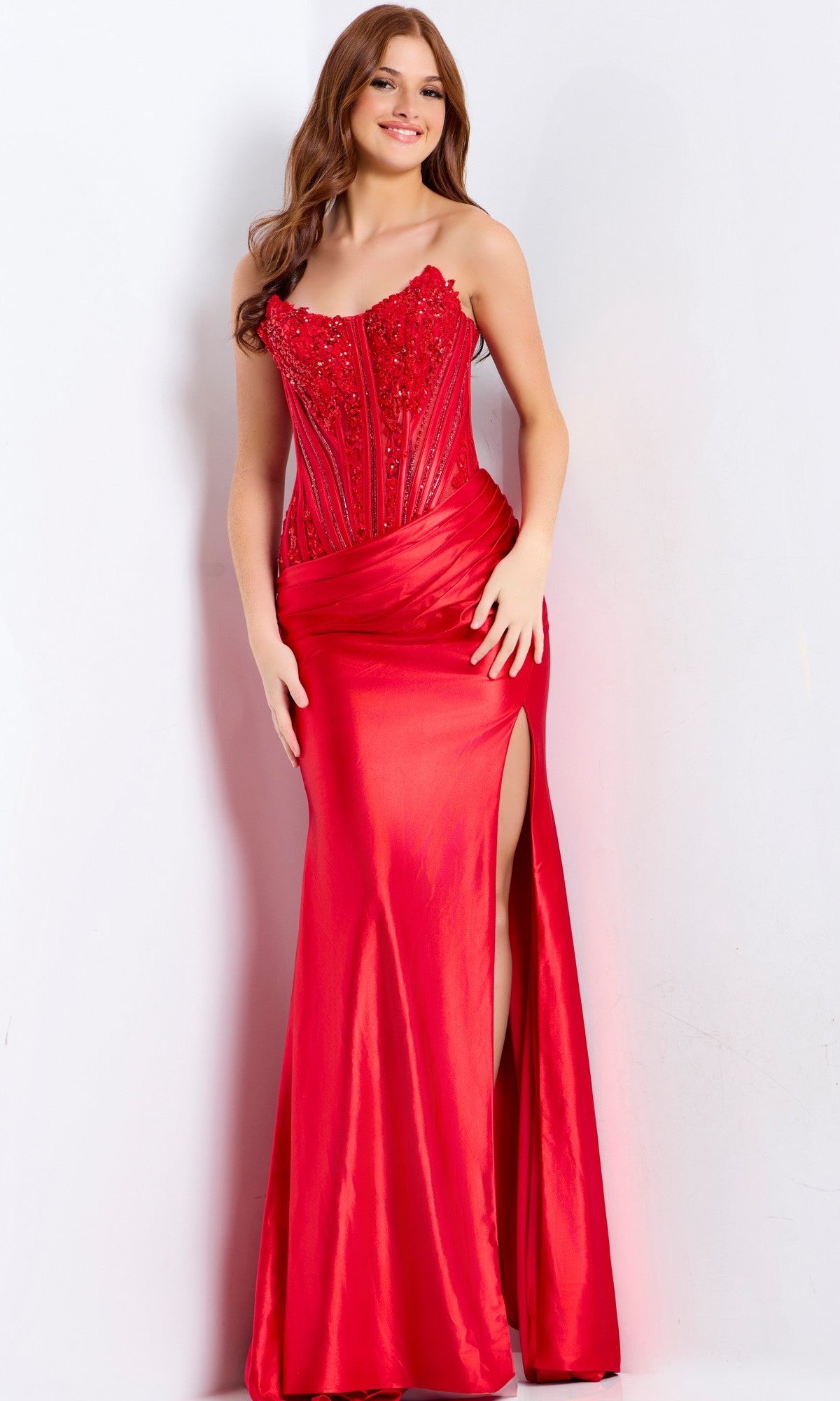 A woman stands against a white background wearing the Jovani 50012 Lace-Corset Strapless Prom Gown in red—a floor-length dress featuring a thigh-high slit and beaded bodice embellishments.